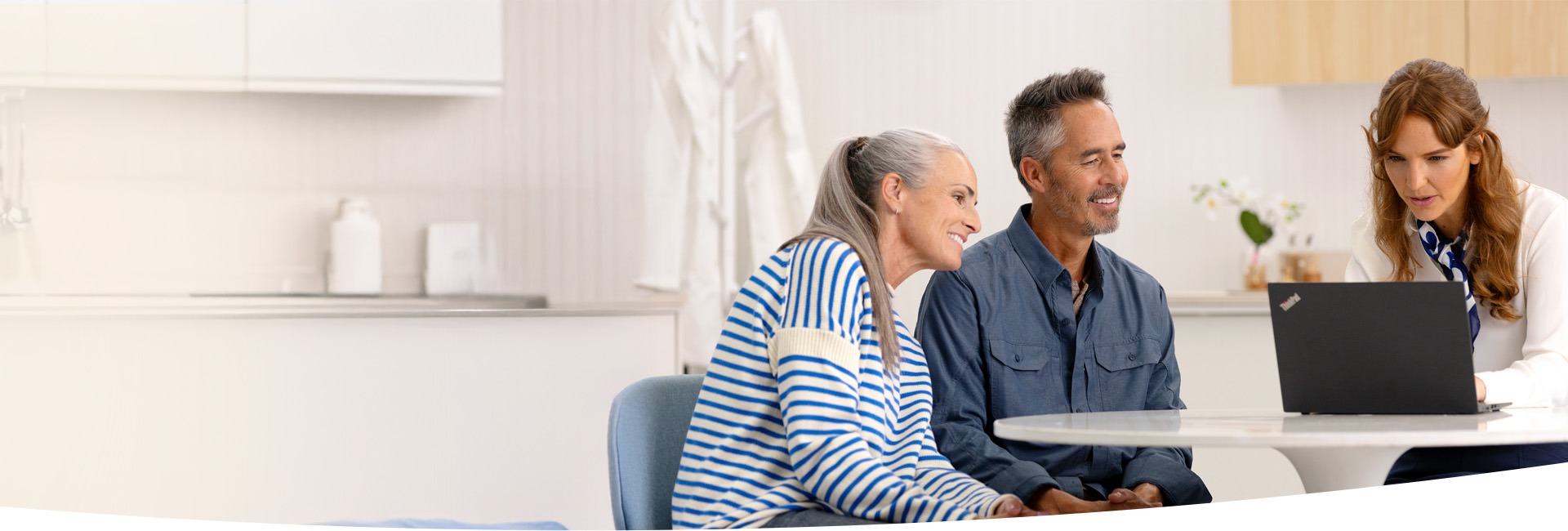  En audionom sitter vid ett bord med en äldre man och kvinna, diskuterar med resulatet av ett hörseltest. Ljust rum med blommor i bakgrunden.