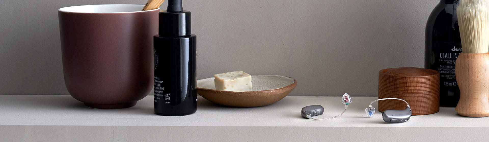  Två gråa hörapparater med avancerad teknik placerade på en hylla omgiven av vardagliga föremål som en kopp, flaska och tvålkopp.