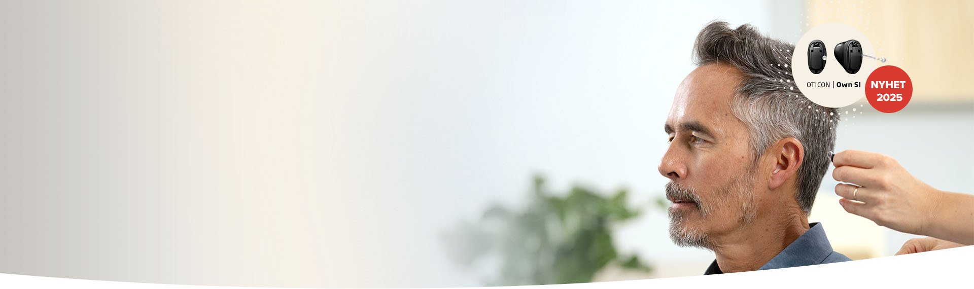  En audionom placerar en Oticon Own SI-hörapparat i en mans öra. Bakgrunden är ljus och suddig med en växt synlig. Text: ’OTICON | Own SI’ och ’NYHET 2025’.