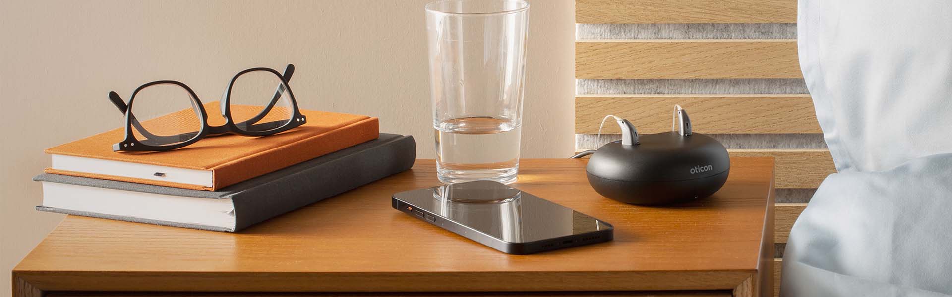  På ett träbord vid sängkanten ligger en bokhög med glasögon ovanpå, ett glas vatten, en smartphone och en Oticon laddningsstation med två hörapparater.
