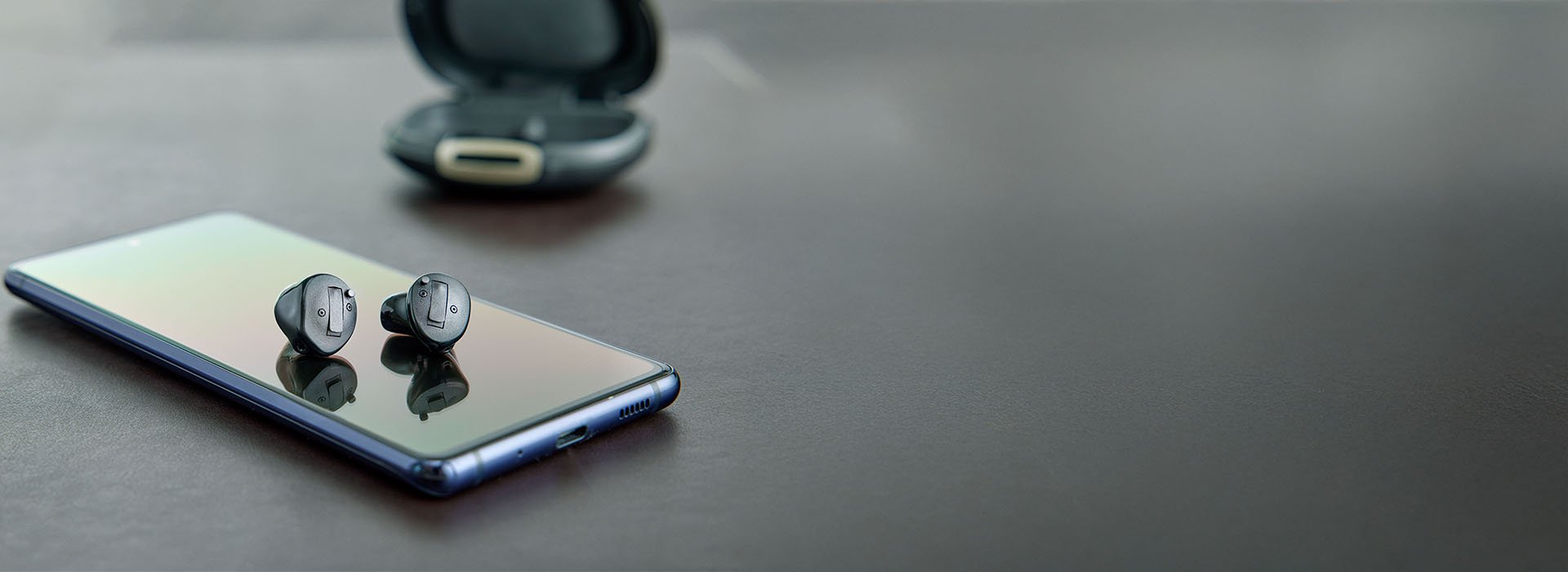  Oticon Own hörapparater placerade på en smartphone med sitt laddningsetui i bakgrunden