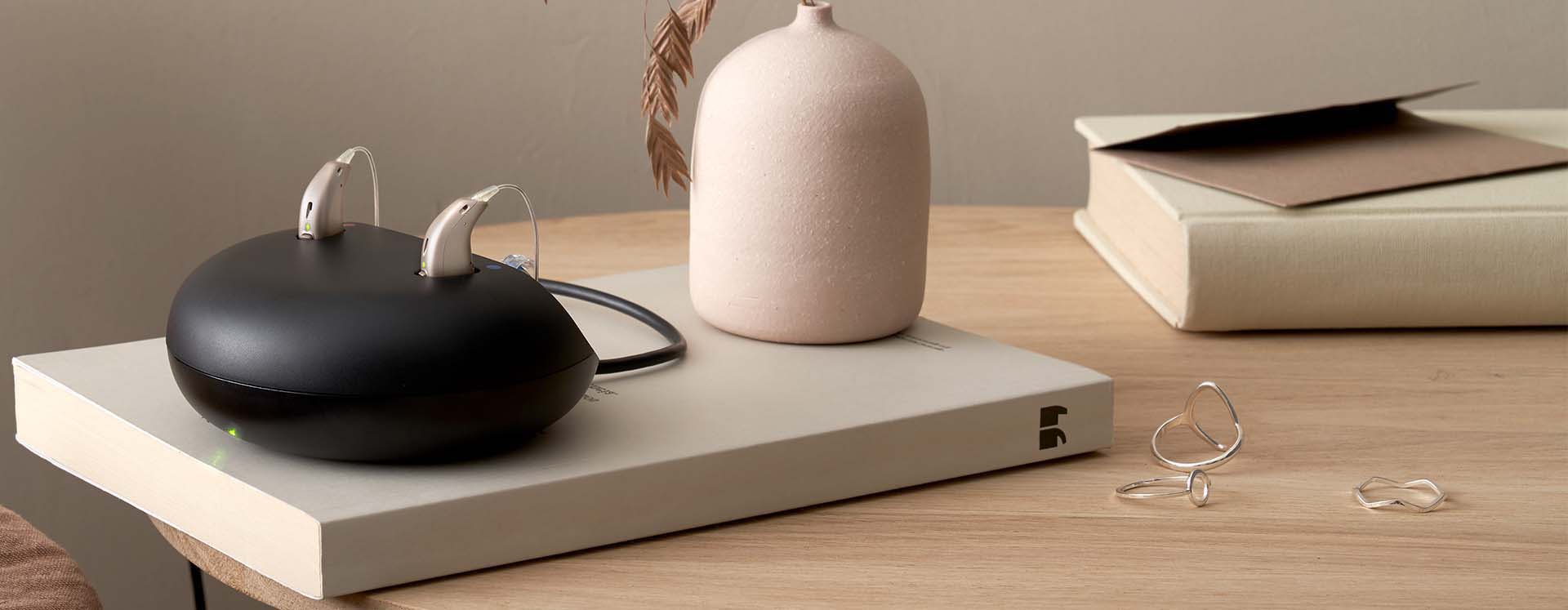  Laddare för uppladdningsbara hörapparater med hörapparater placerade i laddningsstation, på ett bord dekorerat med en bok, smycken och en beige vas.