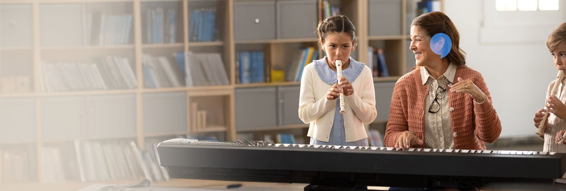  Elektroniskt piano står i förgrunden, medan en lärare med synlig hörapparat från Oticon instruerar två barn som spelar blockflöjt i ett klassrum med bokhyllor i bakgrunden.