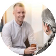  En äldre kvinna med grå fläta som använder hörapparater. Hon sitter i samtal med en man i vit skjorta som håller en kopp. Ljusa, professionella klinikliknande omgivningar.