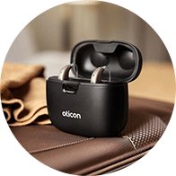 Oticon Real hörapparater i en svart laddningsstation på ett bord. Laddningsbara hörapparater från Oticon för effektiv hörselvård.