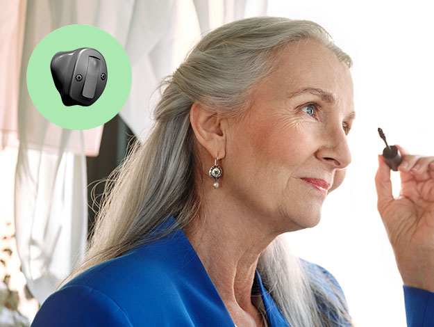  En äldre kvinna med långt, grått hår som har en hörapparat i örat. En i-örat hörapparat från Oticon visas i en grön cirkel.