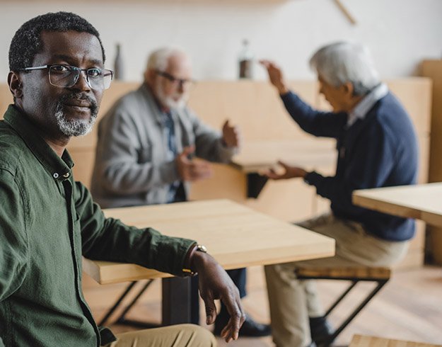  Man som upplever social isolering på grund av obehandlad hörselnedsättning, sittande vid ett bord medan andra två män diskuterar i bakgrunden.
