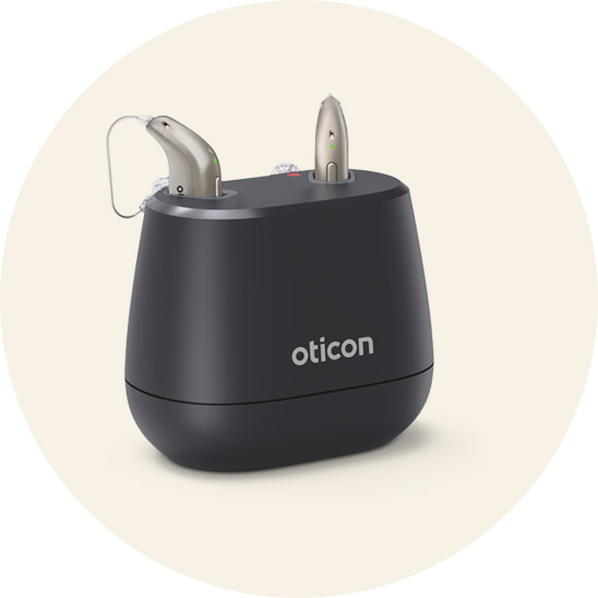  Två silverfärgade Oticon-hörapparater laddas i en svart laddningsstation med Oticon-logotyp. Produkten är avsedd för hörselvård och användarvänlighet.