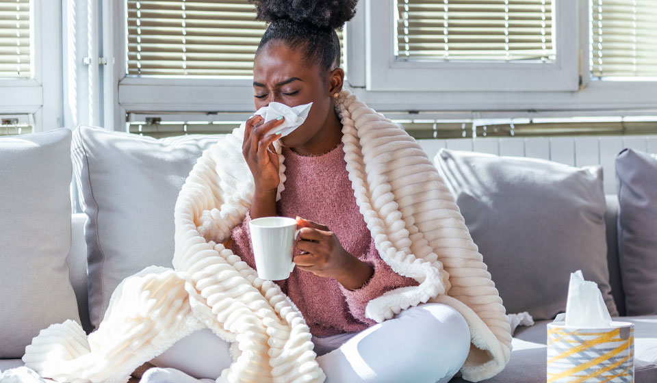 Woman wrapped in a blanket sitting on a couch with a cup of tea, tissues nearby, suggesting she has a cold.