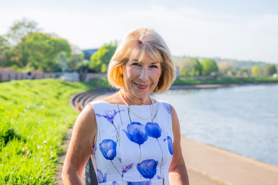 Jennie Bond wearing a floral dress standing near a scenic riverside walkway on a bright day.