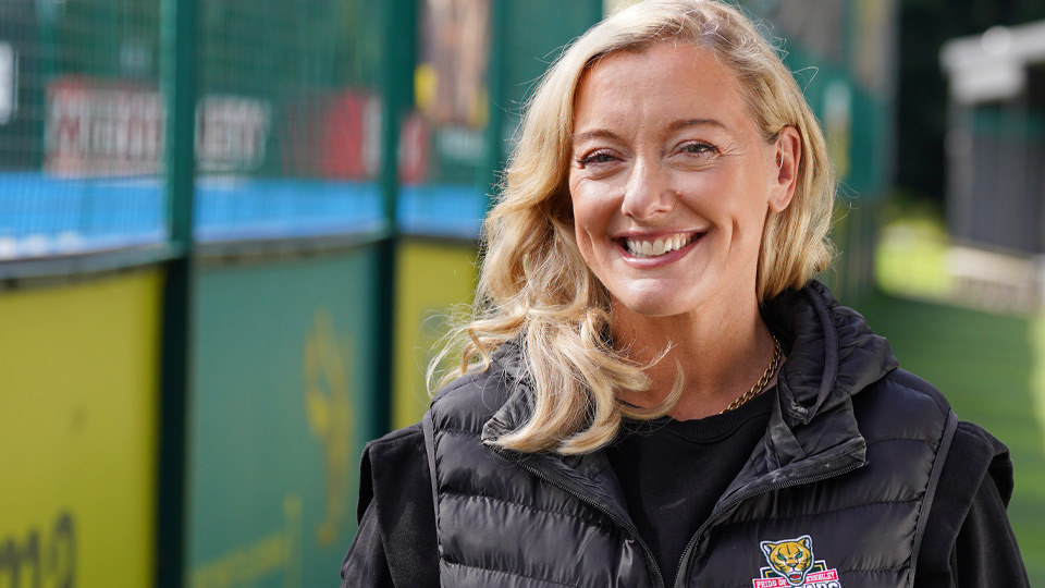 Blonde woman outdoors wearing a black puffer vest with a colorful tiger logo, standing near a fenced sports area.