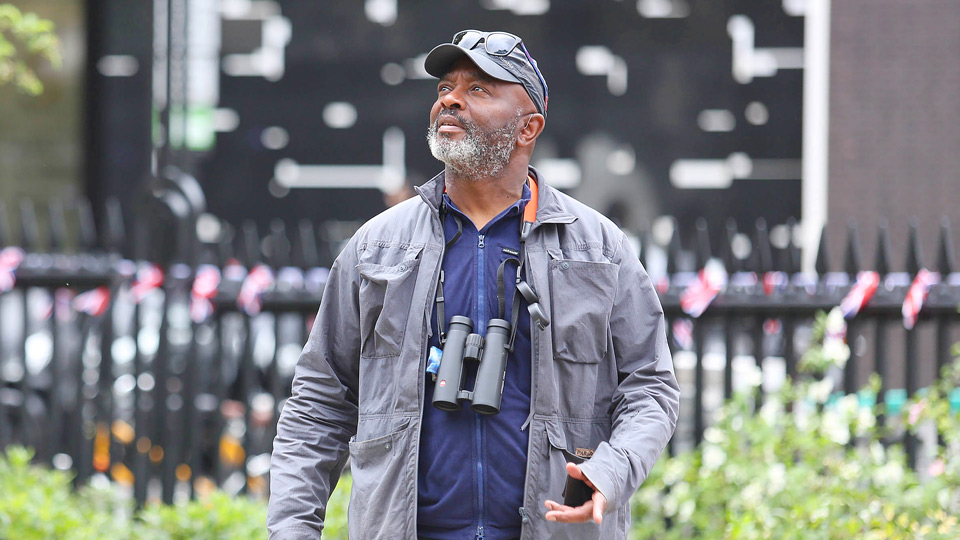 Man outdoors holding a smartphone, wearing binoculars around his neck, with a blurred urban background.