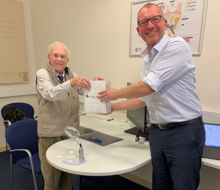 Hearing care professional handing a gift bag to an elderly man as part of a give-back program in a clinic setting.
