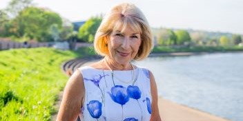 Smiling older woman standing outdoors near a waterfront path, with greenery and sunlight in the background.