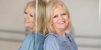 Senior woman in a blue blouse leaning against a mirror, reflecting on hearing health.