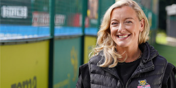 Blonde woman outdoors wearing a black jacket near a fenced area.