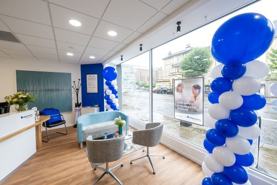 Modern interior of Audika branch in Bristol, featuring blue and white balloon decorations, a reception desk, seating area, wooden flooring, and large windows overlooking the street.