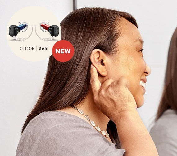 A person adjusts an in-ear hearing aid in a modern indoor setting. The image highlights ’Oticon Zeal’ hearing aids with a ’NEW’ label, showcasing discreet, high-performance hearing solutions.