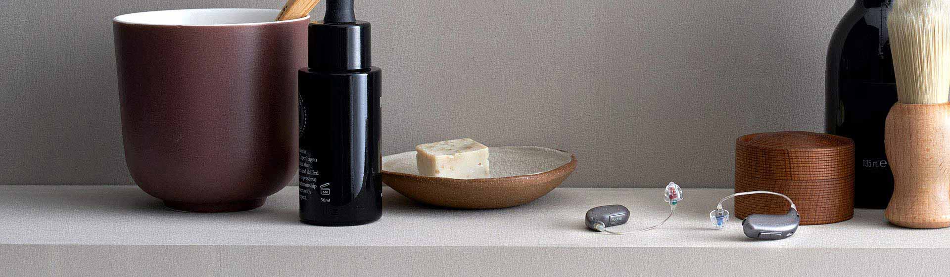 Digital hearing aids displayed on a neutral surface alongside grooming accessories and a soap dish.