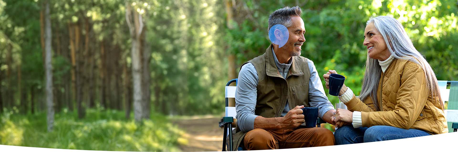 Older couple sitting outdoors on a bench in a forest, sharing a conversation while holding mugs.