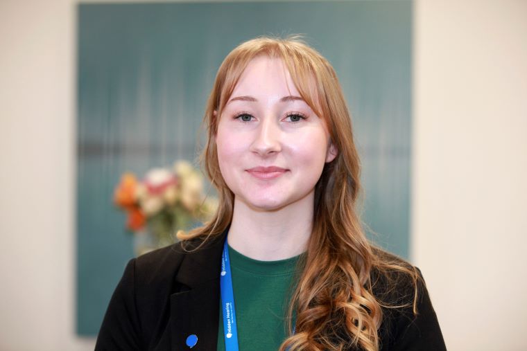 Audika audiologist wearing a badge, standing in front of a teal background with blurred flowers.