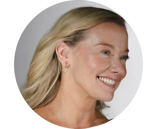 A close-up side view of a woman's ear with blonde hair, showing a discreet behind-the-ear hearing aid in a minimalist, professional setting.