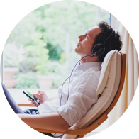 Man sitting on a chair near a window, wearing headphones and holding a smartphone, enjoying music in a serene setting.