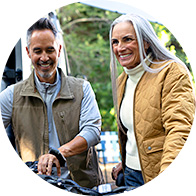 Man in a brown vest and woman in a mustard quilted jacket standing outdoors next to a bicycle surrounded by greenery in a casual setting.