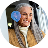 Woman with long gray hair smiling, wearing a yellow quilted jacket, with a highlighted blue ear, promoting hearing health awareness.