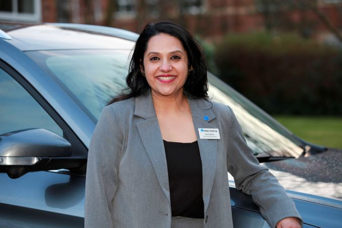 Audika audiologist standing outdoors beside a car, wearing a professional suit and name badge.