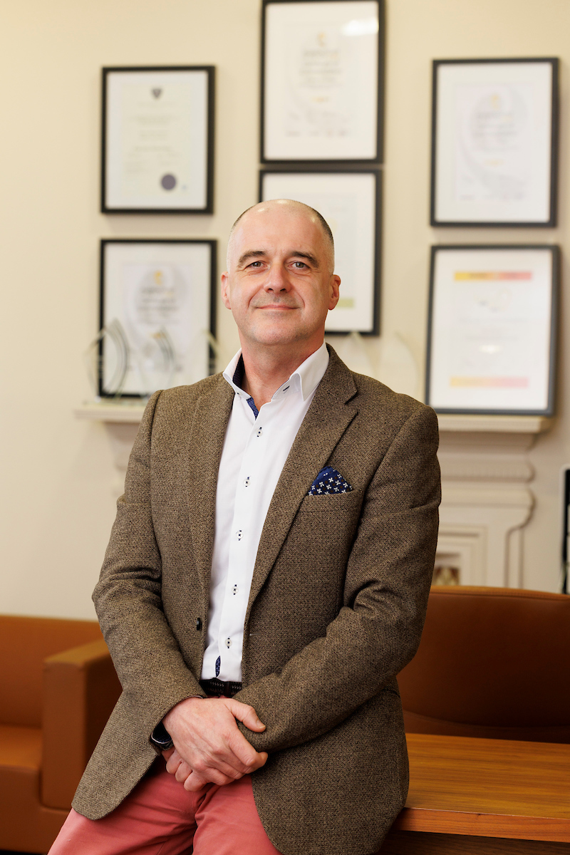 A professionally dressed individual sits with crossed hands in a hearing clinic office, surrounded by framed certificates and awards on the wall, reflecting expertise in audiology services.