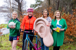Four people are outdoors near a lake and trees. Two hold sports items (rugby ball, bike), a woman holds a large ear model, and another holds a book titled 'Hidden Hearing.'