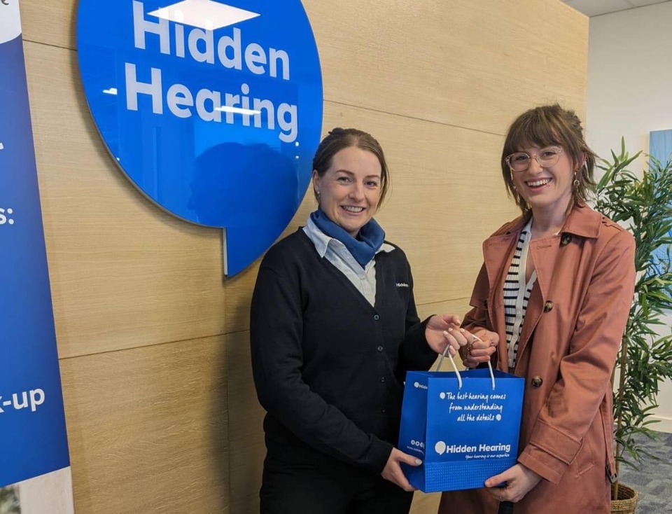Two individuals stand inside a hearing clinic beside a blue 'Hidden Hearing' logo on a wooden wall. One holds a branded gift bag with the text: 'The best hearing comes from understanding all the details.' The setting features indoor greenery and informational banners.