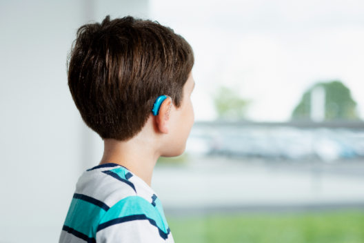 A young boy wearing a blue hearing aid looks out a window, with a blurred outdoor scene of greenery and parked cars in the background, suggesting a hearing clinic setting.