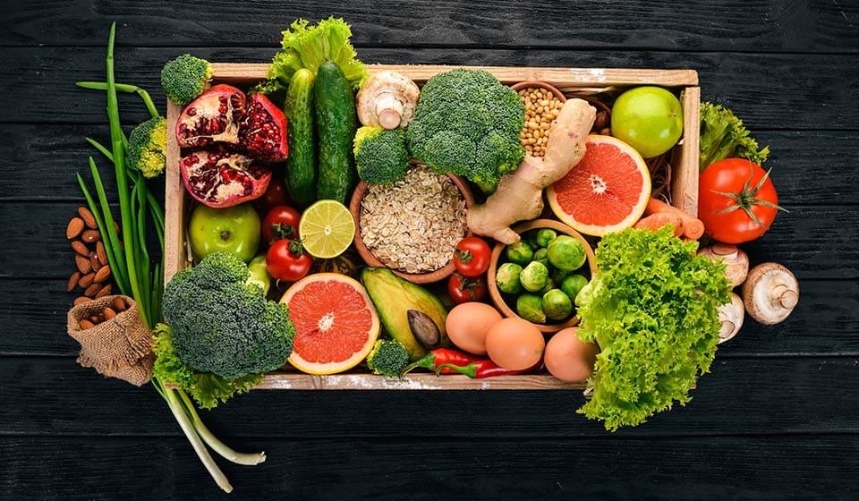 Una cassetta di legno contiene vari alimenti freschi: broccoli, avocado, pomodori, lattuga, melograno, pompelmo, uova, avena e funghi, disposti su un tavolo nero.