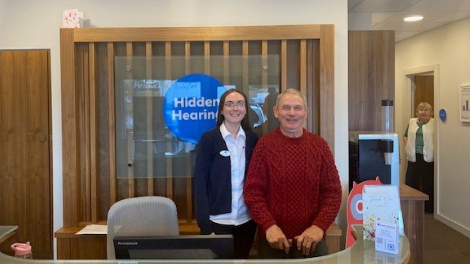 A hearing clinic reception area with a 'Hidden Hearing' logo prominently displayed on a blue circular sign. A professional staff member stands alongside a visitor near a desk with a computer monitor, brochures, and a 'Thank You' card. Surroundings include wood-panel decor, a coffee machine, and an open hallway.