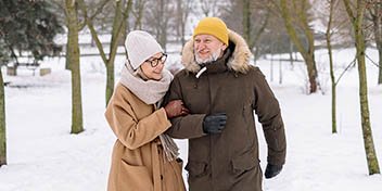 [GEN] A couple walks arm-in-arm through a snowy park, dressed in winter coats, hats, and scarves. Leafless trees and a white, snow-covered landscape form the serene background. No text visible.