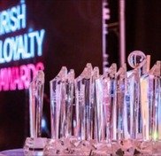 [GEN] A row of crystal trophies is displayed on a table, illuminated by vibrant pink and blue lights. In the background, partially visible text reads, 'IRISH LOYALTY AWARDS,' suggesting an award ceremony context.