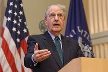 A suited figure stands at a podium, gesturing with one hand, framed by U.S. and institutional flags in a formal setting, suggesting a speech or presentation in a professional environment.