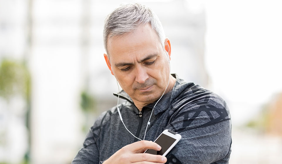 Hombre con cabello canoso usando auriculares conectados a un teléfono móvil durante una actividad al aire libre.