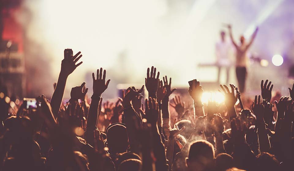 People raising hands at a lively concert with bright stage lights in the background.
