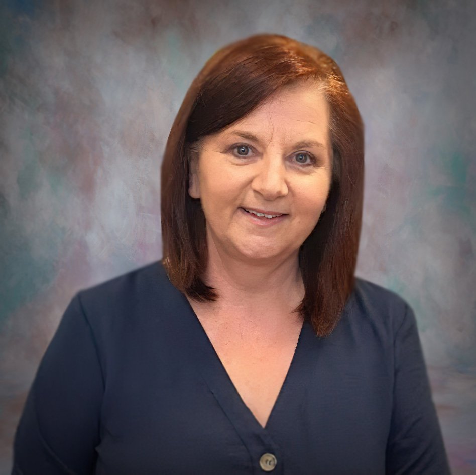 A person with shoulder-length auburn hair wears a navy buttoned blouse against a softly textured, pastel-colored studio backdrop. The face is purposely blurred to maintain anonymity. No text or hearing aids are visible.