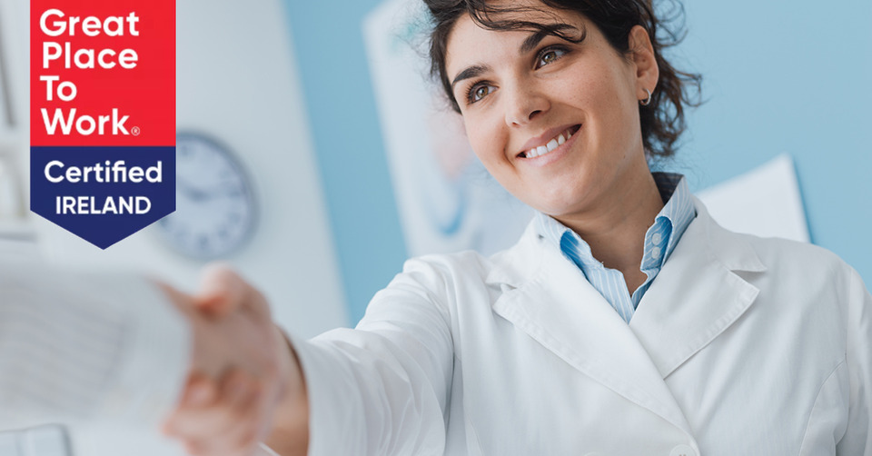 A healthcare professional in a white coat extends a document or leaflet in a clinical setting with blue walls. Text reads, 'Great Place To Work Certified IRELAND.' Context implies a hearing clinic.