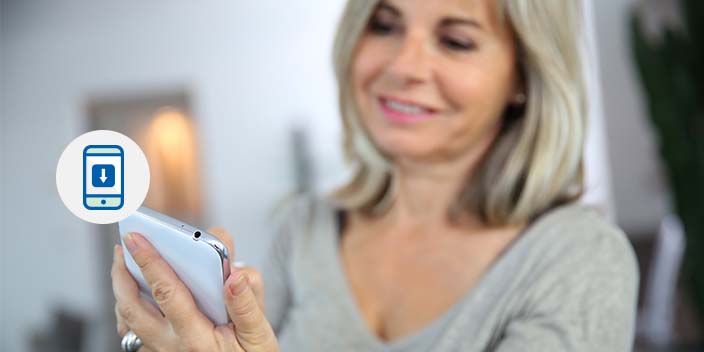 A woman with shoulder-length blonde hair holds a smartphone in a modern indoor setting. A graphic overlay depicts a phone icon with a download symbol, emphasizing mobile app functionality, possibly for hearing aid management. 