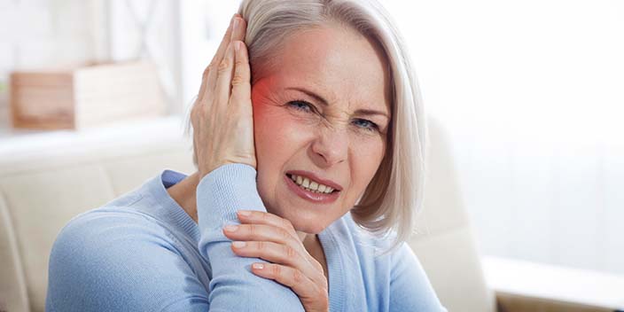 [GEN] A senior woman with silver hair, wearing a light blue sweater, touches her ear while seated on a beige sofa in a softly lit living room. No text is visible.