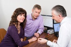 [GEN] A hearing care professional discusses hearing aid options with a man and woman at a desk in a hearing clinic. A computer displaying hearing test results and hearing devices are visible.