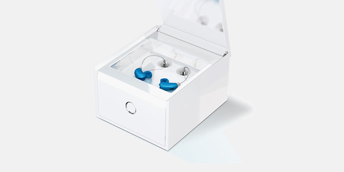A sleek white charging case holds two modern hearing aids with blue ear molds, positioned open on a clean, white background.