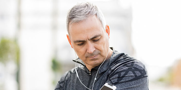 A silver-haired individual wearing a dark athletic jacket listens to wired earphones connected to a device, standing outdoors in a bright urban environment with blurred greenery and structures. No text visible.