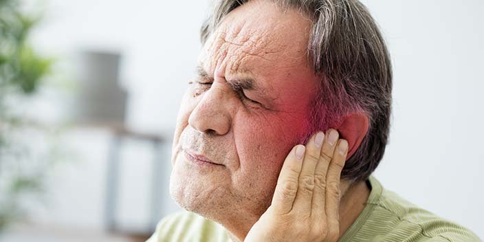 Una persona sostiene su mano cerca de la oreja izquierda, posiblemente indicando incomodidad o problemas auditivos. El fondo es un espacio interior difuminado con muebles y plantas visibles.
