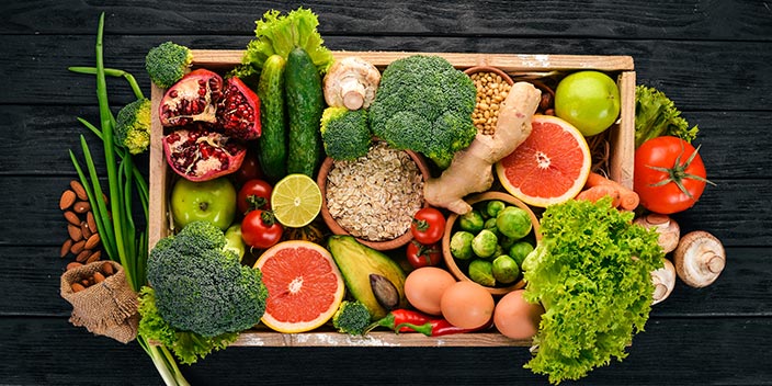 Una caja de madera contiene frutas, verduras y alimentos saludables como brócoli, tomates, aguacate, granada, jengibre, huevos, avena, lechuga y limón, sobre una mesa negra.